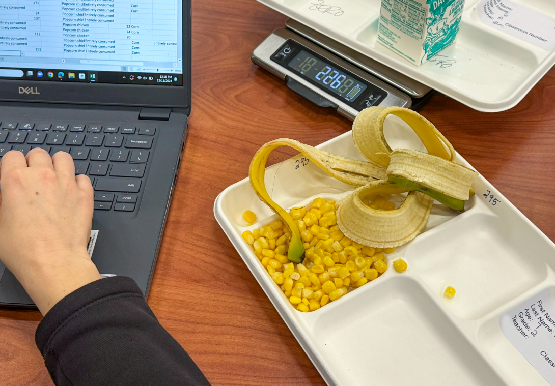 A researcher weighs a carton of milk and records the data on a laptop. A school lunch tray with corn and a banana peel is in the foreground.