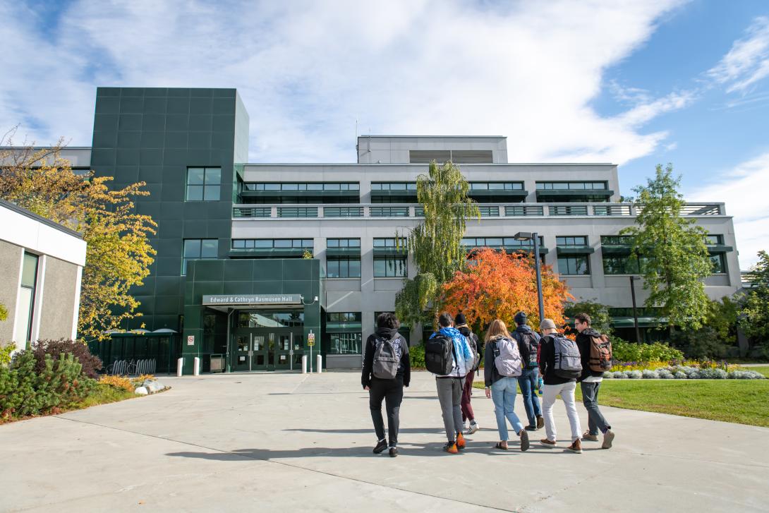 A group of students walks toward the main entrance of Rasmuson Hall. The trees are starting to turn orange.