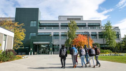 A group of students walks toward the main entrance of Rasmuson Hall. The trees are starting to turn orange.